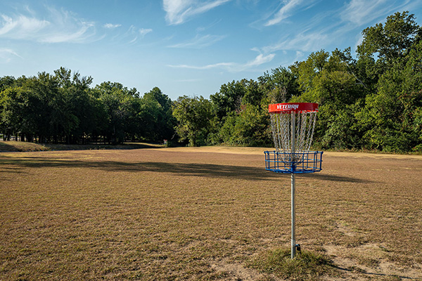 A metal disc golf basket stands on a dry, grassy field with trees and a partly cloudy sky in the background.