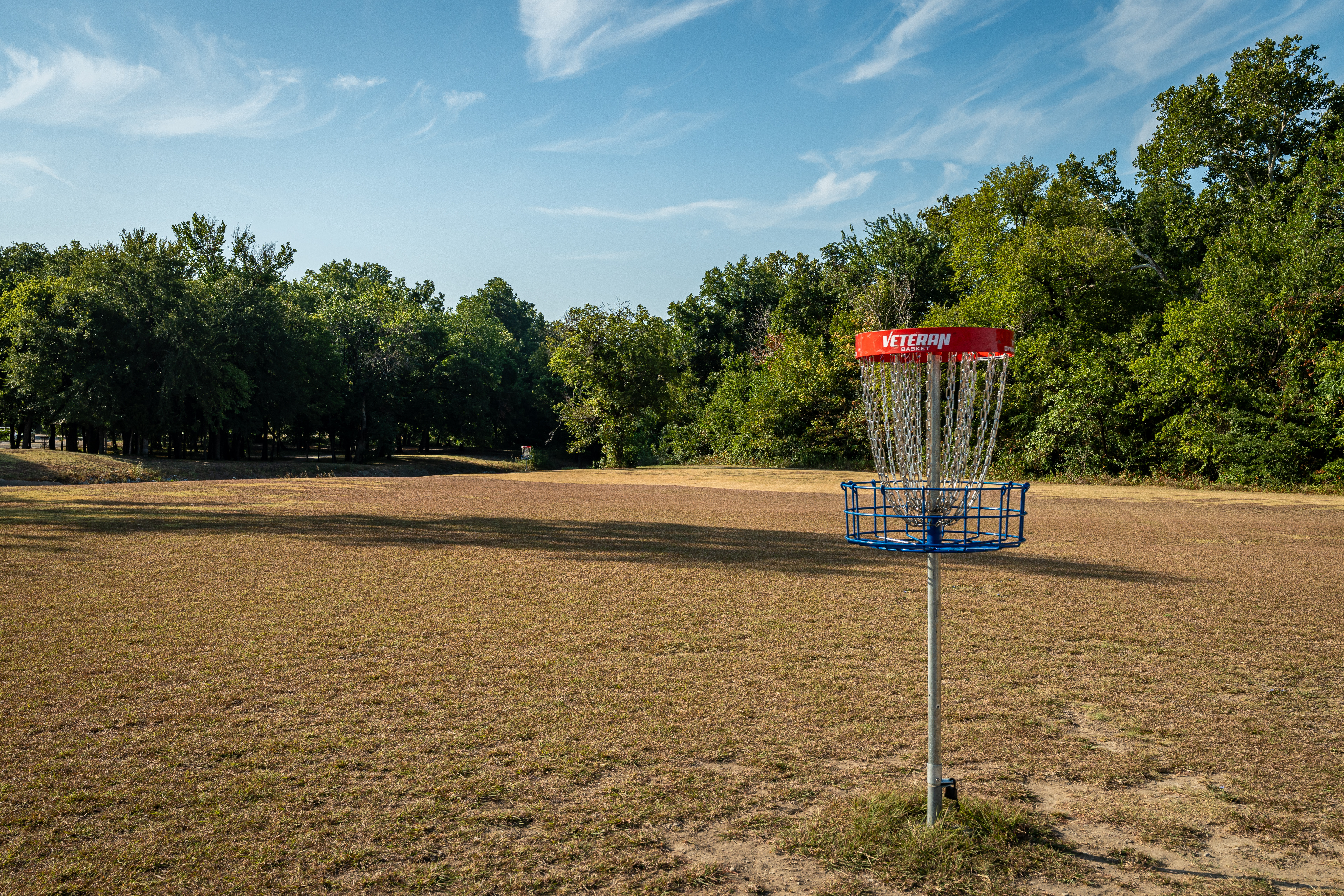 Disc golf basket in the middle a field with dense trees in the background, Alex Clark Memorial Disc Golf Course, McKinney