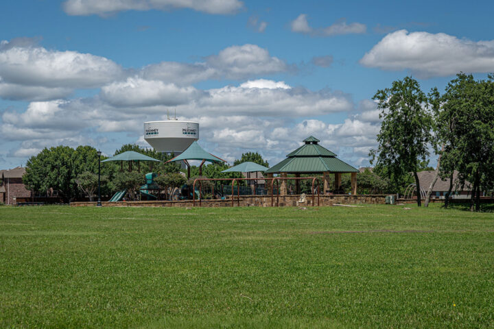 A park with green grass, playground equipment, and covered picnic areas; trees and a water tower are visible in the background under a partly cloudy sky.