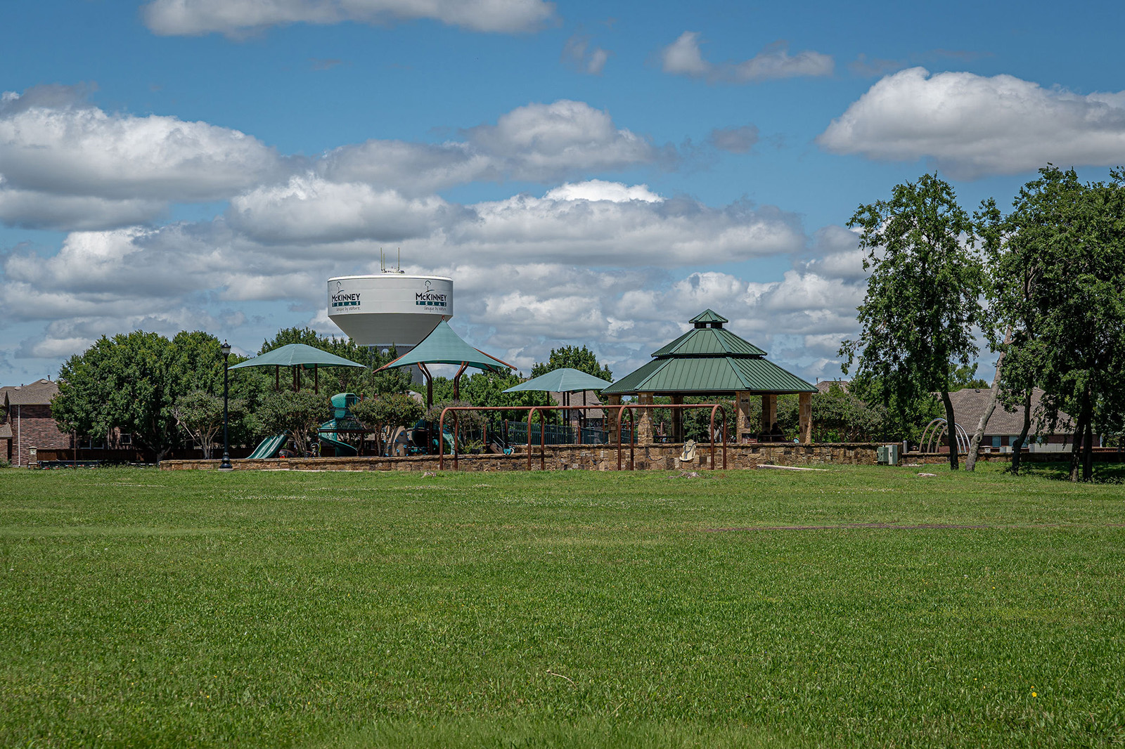 A park with green grass, playground equipment, and covered picnic areas; trees and a water tower are visible in the background under a partly cloudy sky.