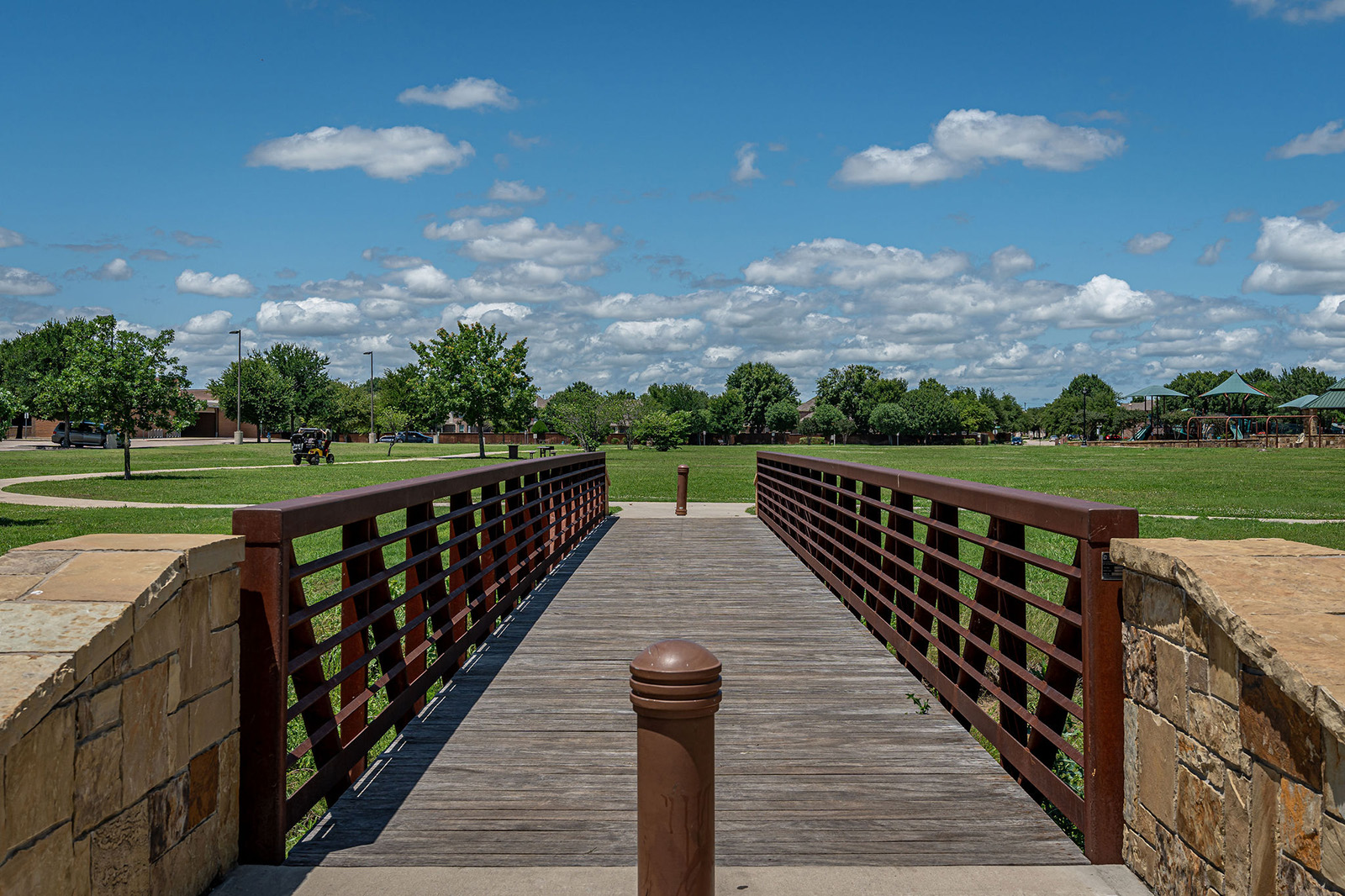 A wooden footbridge with metal railings leads to a grassy park area with trees, benches, and a partly cloudy sky overhead.