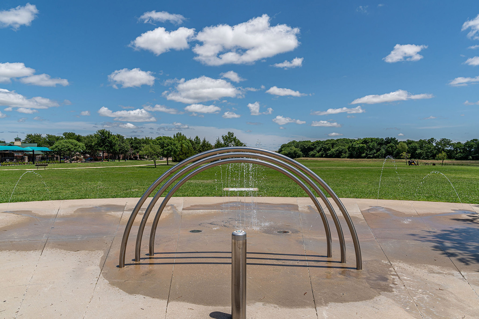 A splash pad with metal arches spraying water, set in a park with green grass, trees, and a blue sky with scattered clouds.