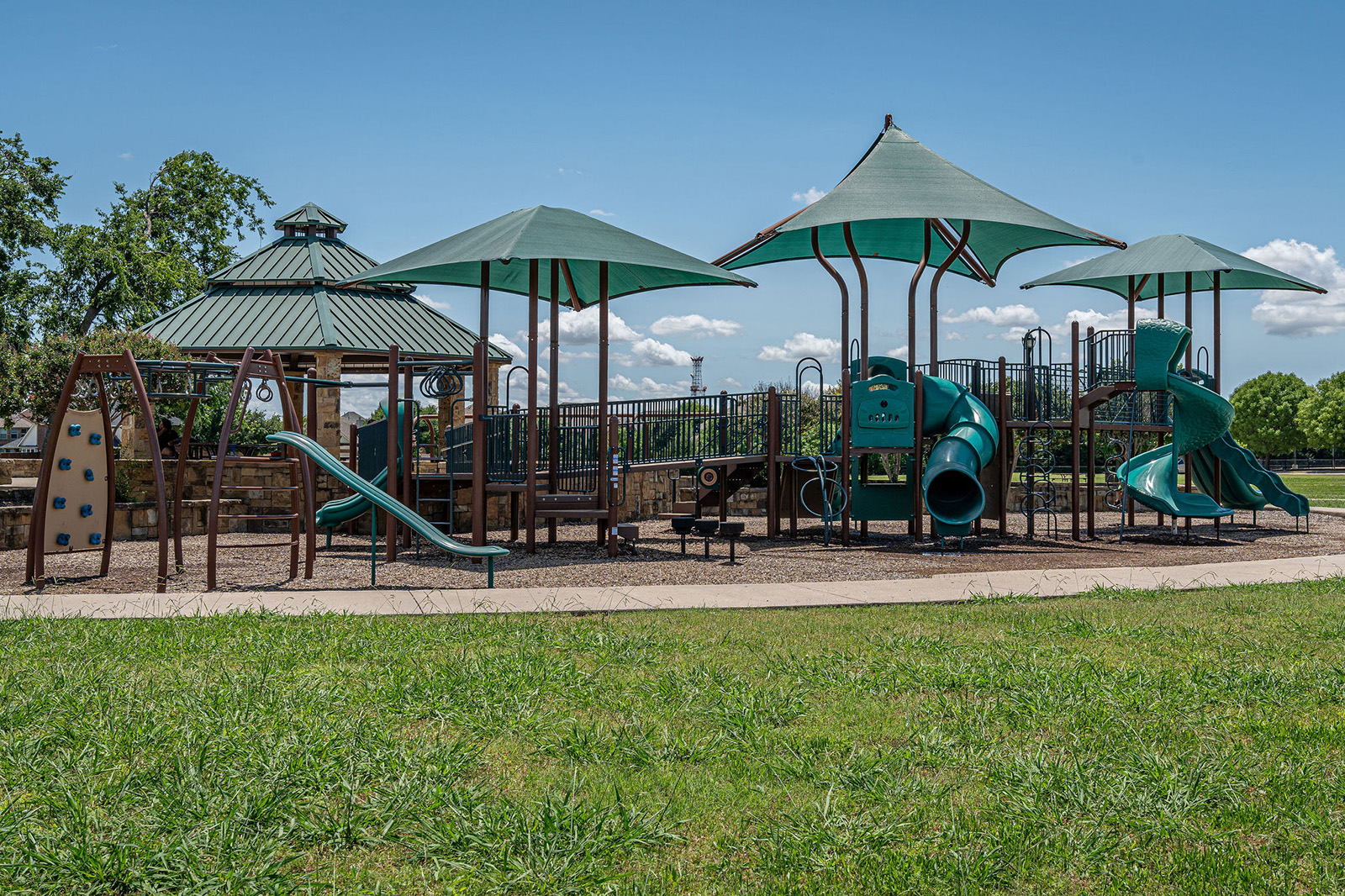 A playground with slides, climbing structures, and shade canopies on a mulch surface, surrounded by grass and trees under a clear blue sky.