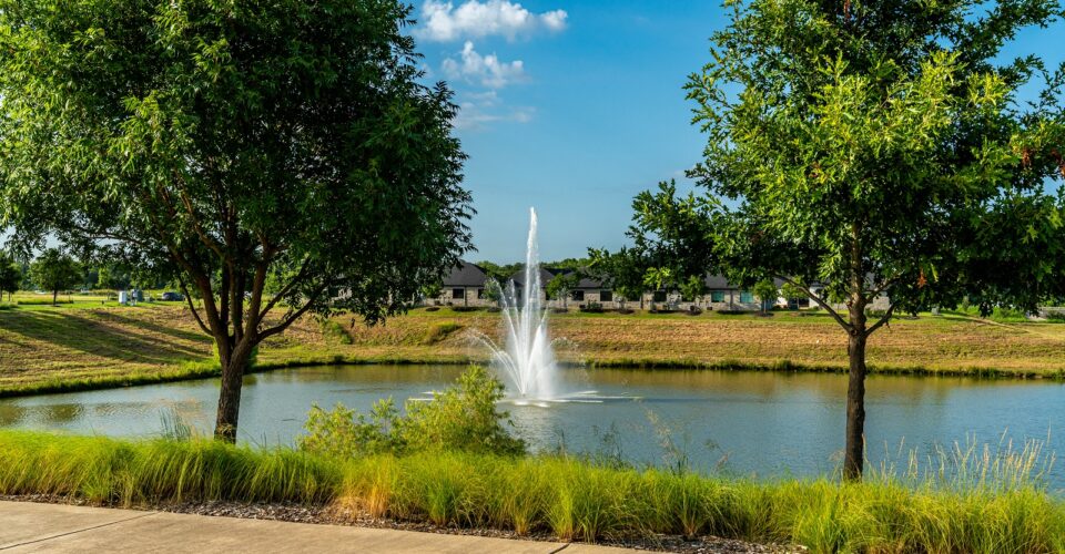 Auburn Hills trail concrete path with landscaping and green grass, and a beautiful water fountain in the middle of the pond