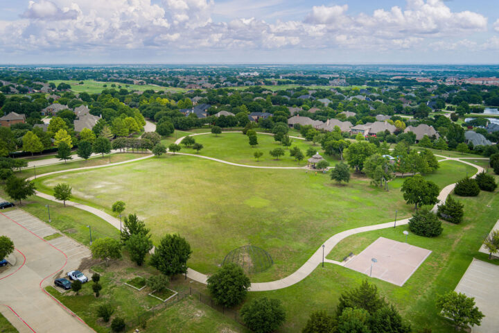 Aerial view of a suburban park with open green space, walking paths, scattered trees, a gazebo, and a basketball court, surrounded by residential homes.