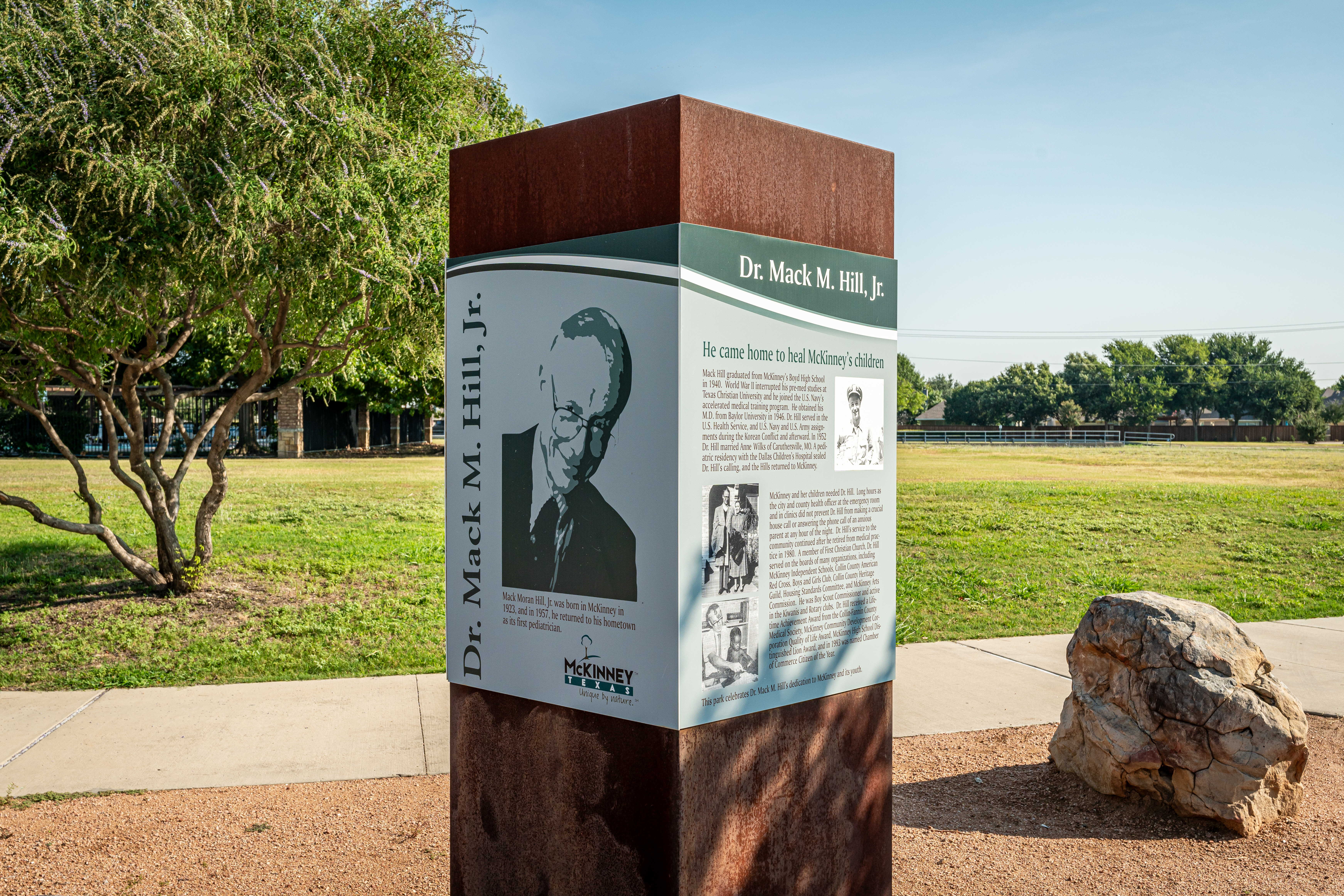 A monument depicting Dr. Mack M. Hill, Jr. stands in a sunny park. The sign features a portrait and text about his contributions. Nearby are a tree and a large rock.