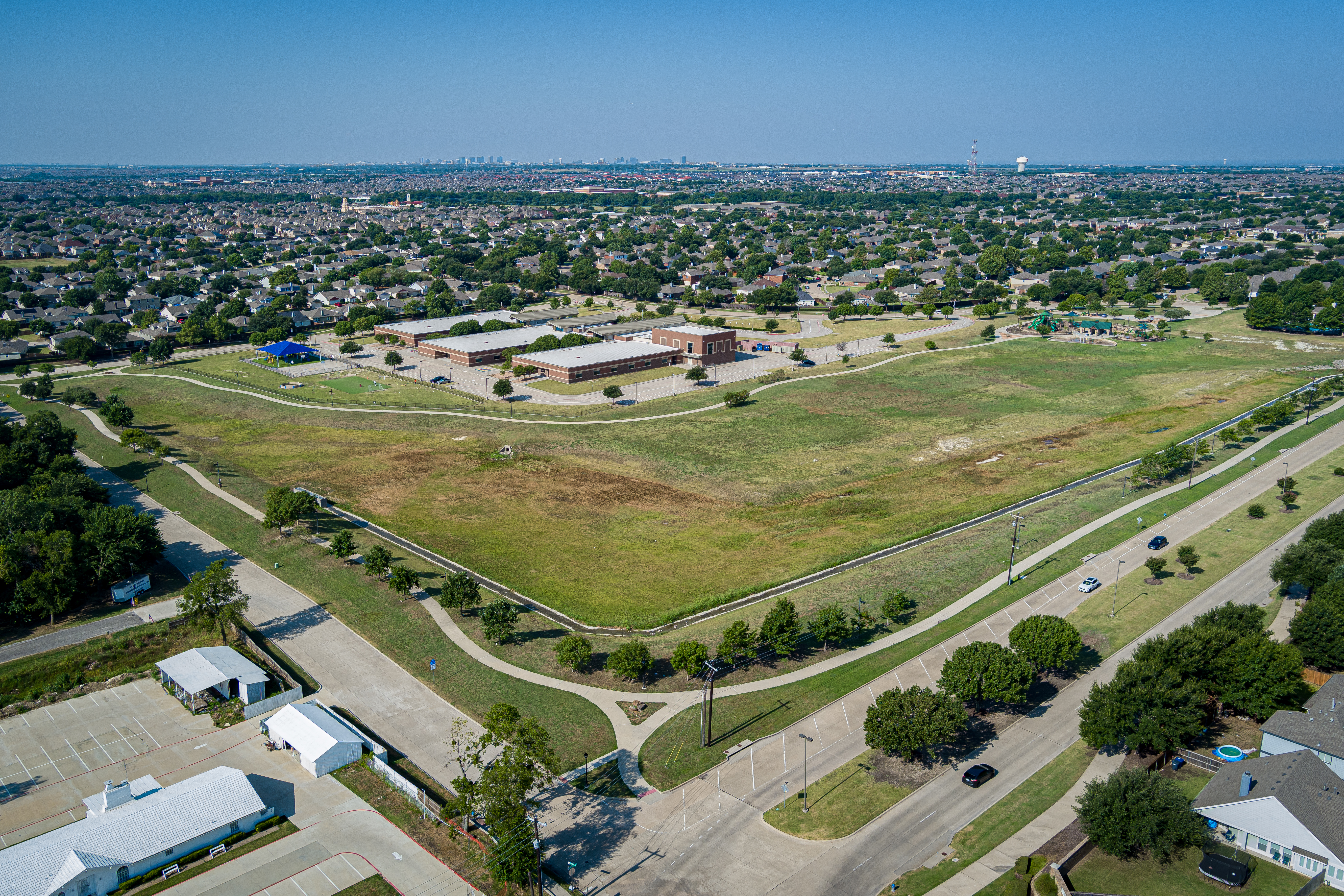 Aerial view of a large grassy field bordered by a suburban neighborhood. Houses and trees surround a central building complex under a clear blue sky.