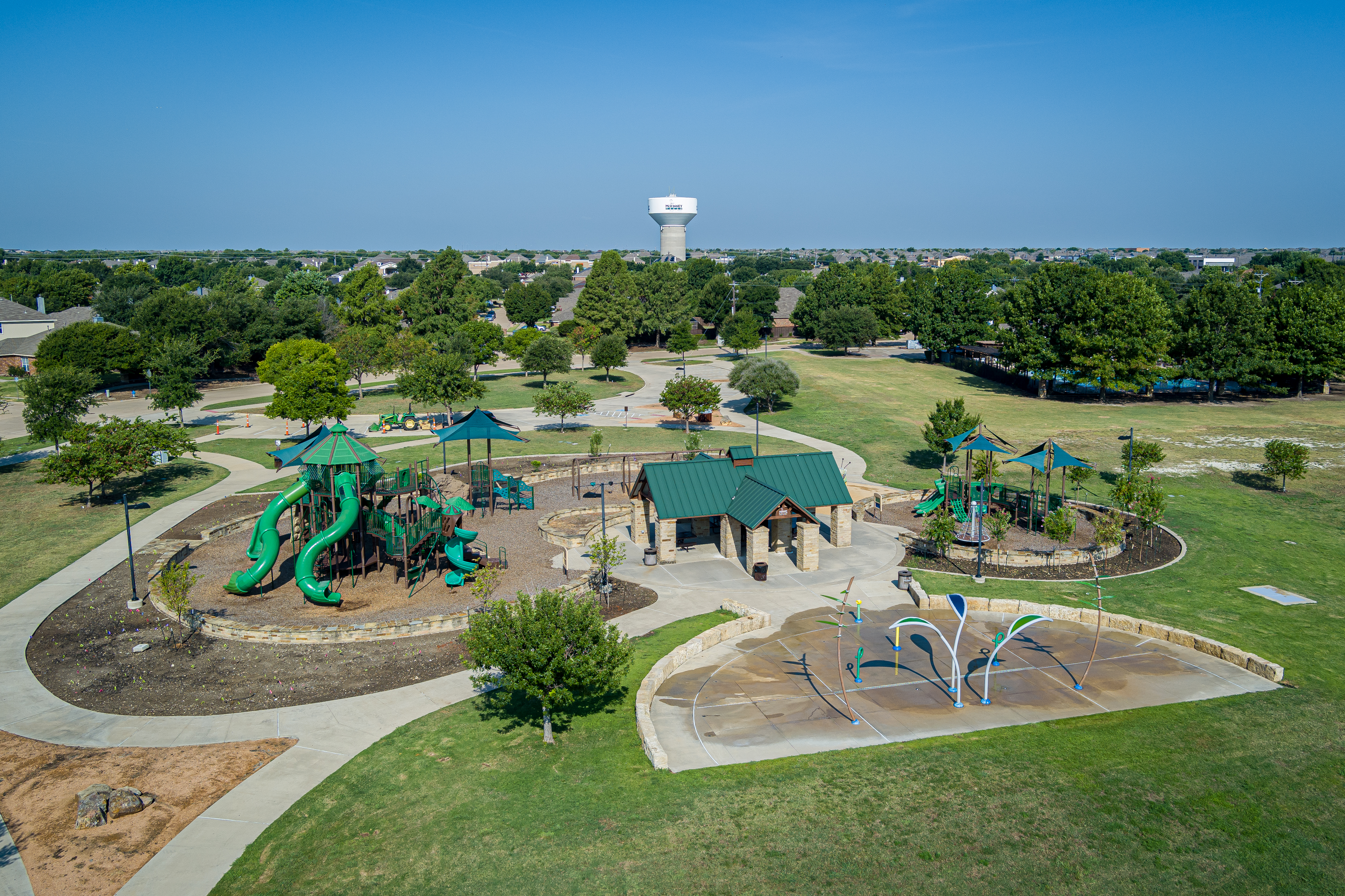 Aerial view of a park with green playground equipment, a pavilion, and a splash pad. Surrounded by walking paths, trees, and open grassy areas under a clear blue sky.