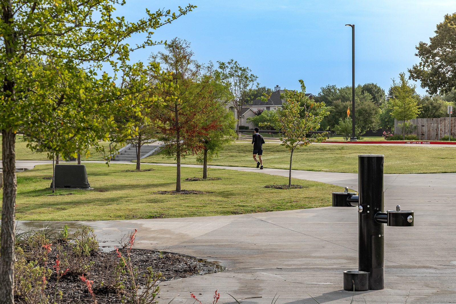 E.A. Randles park show of the walking trail loop with a water fountain in foreground and man running in the background with trees all around.