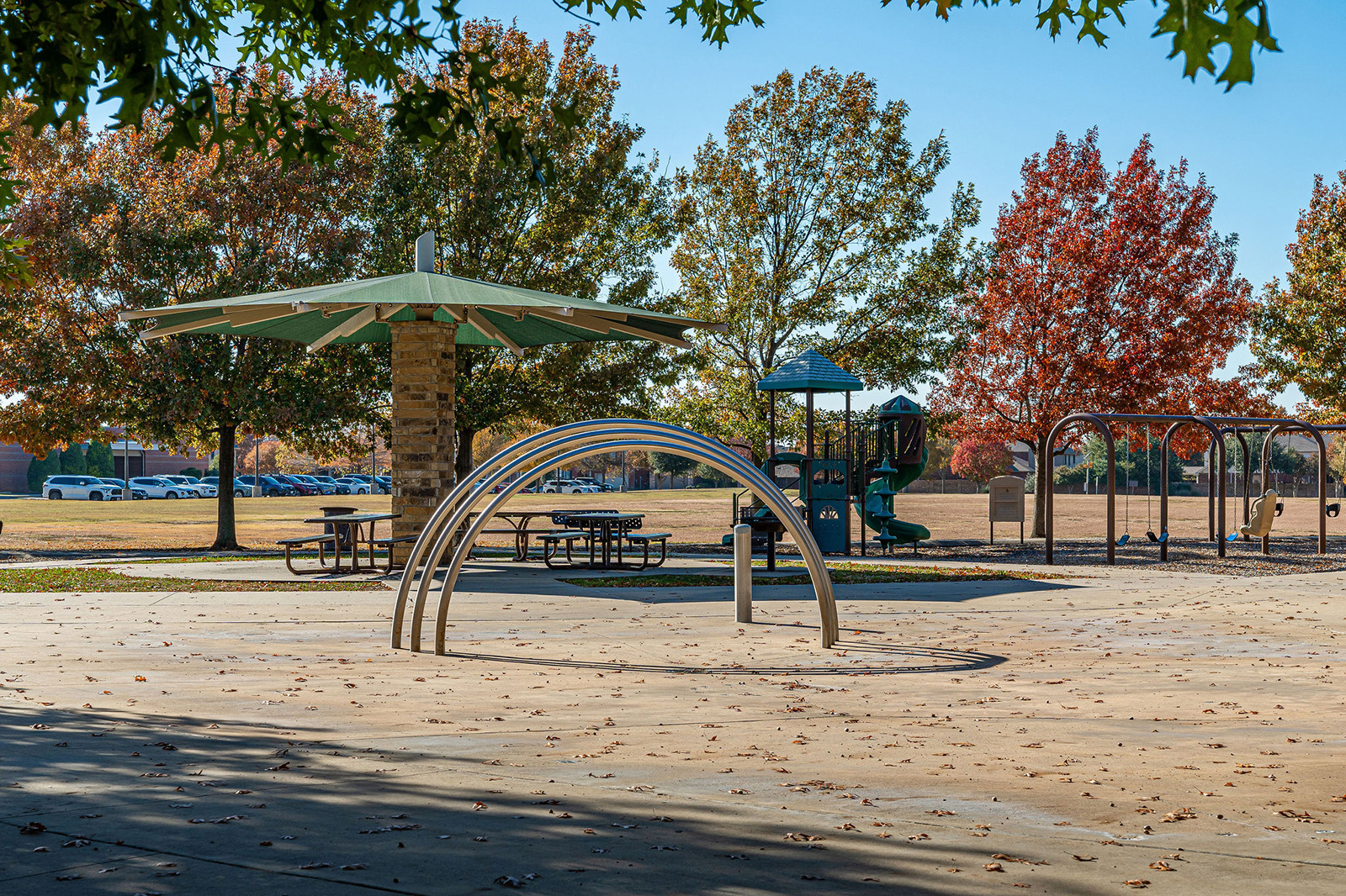 A playground with metal climbing arches, picnic tables under a pavilion, swings, and trees with autumn foliage in the background.