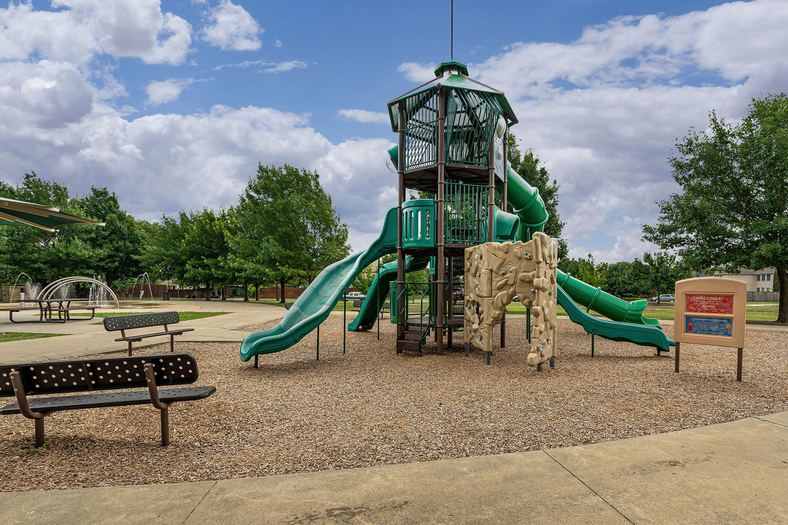 A playground with green slides, a climbing wall, and a covered tower structure sits on a bed of wood chips, with benches and trees nearby under a partly cloudy sky.