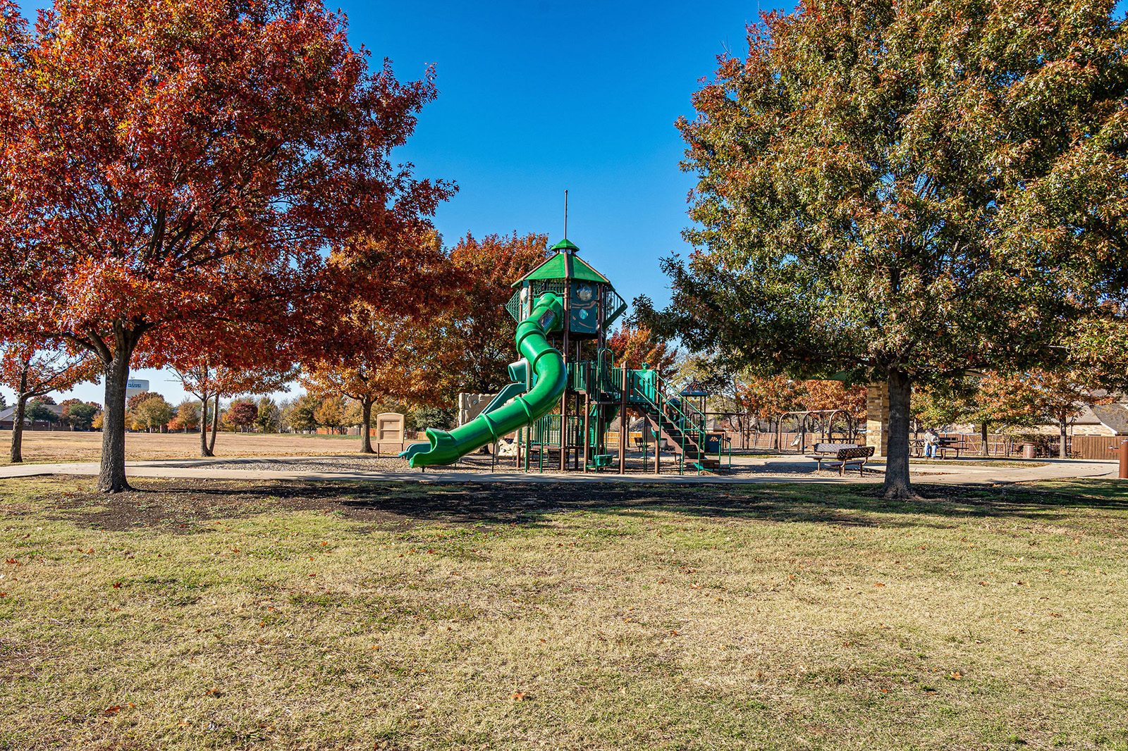 A playground with a large green spiral slide and climbing structure surrounded by autumn trees in a grassy park.