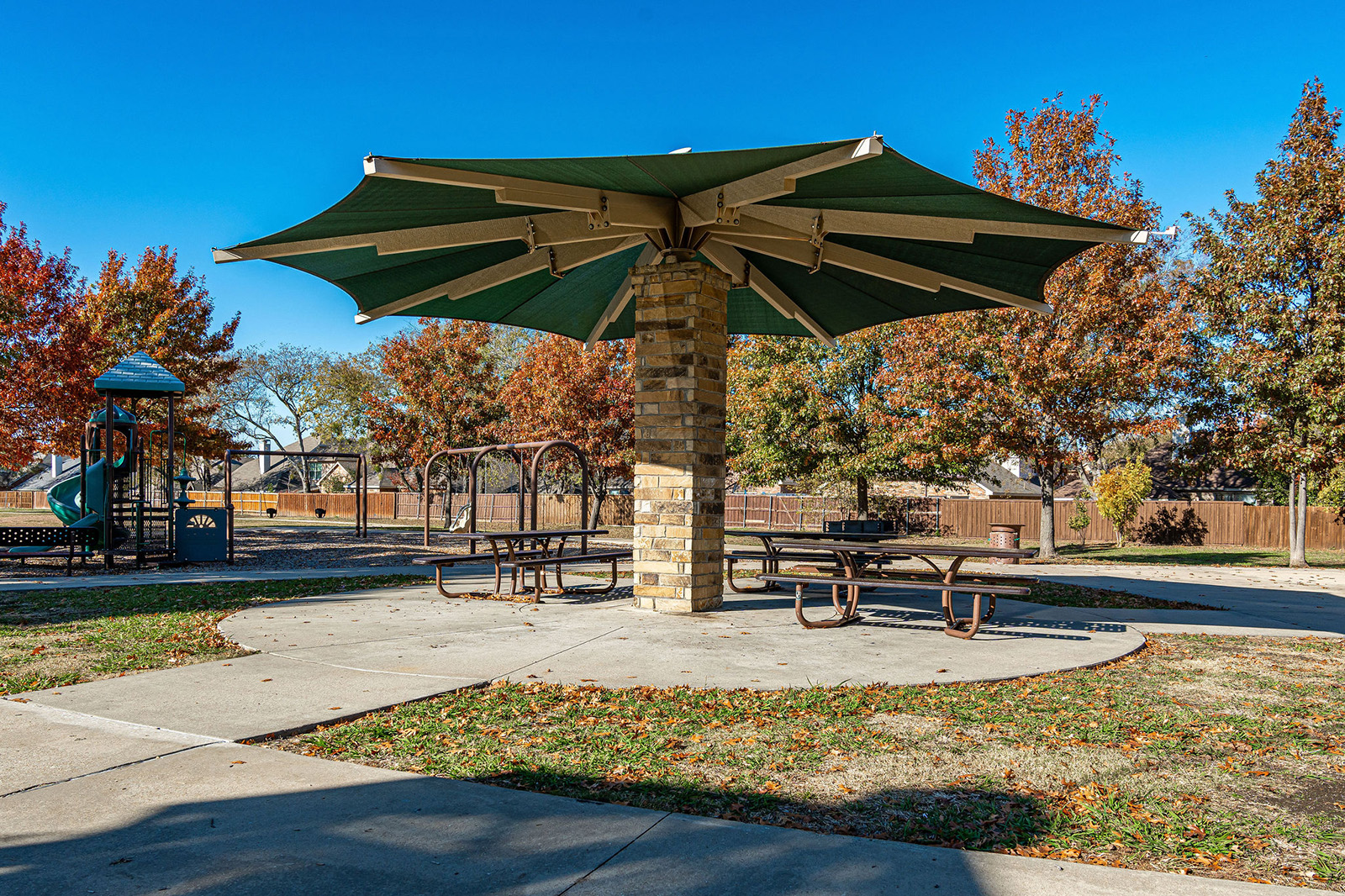 A large green umbrella canopy shades picnic tables on a concrete area in a park with trees and a playground in the background.