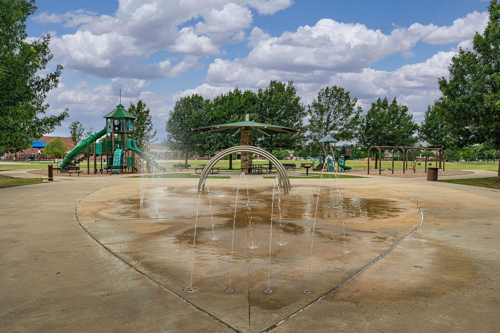 A playground with green play structures, a splash pad with water jets in the foreground, trees, and a partly cloudy sky.