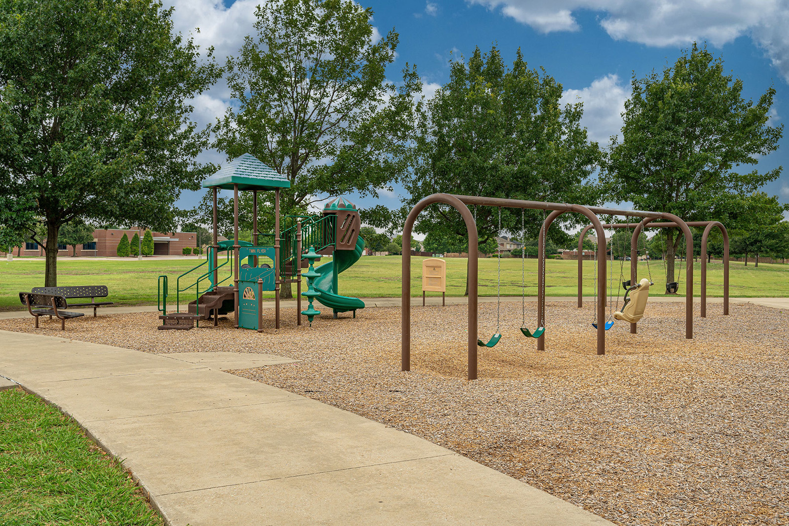 Outdoor playground with a slide, climbing structure, swings, bench, and mulch ground covering, surrounded by grass and trees under a partly cloudy sky.