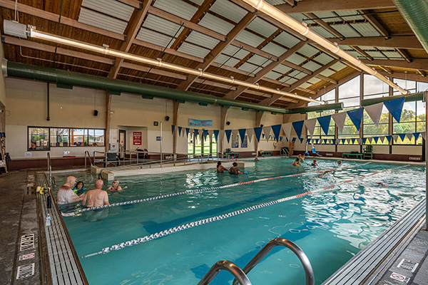 Indoor swimming pool with several people in the water, separated by lane markers, under a wooden ceiling with large windows letting in natural light.