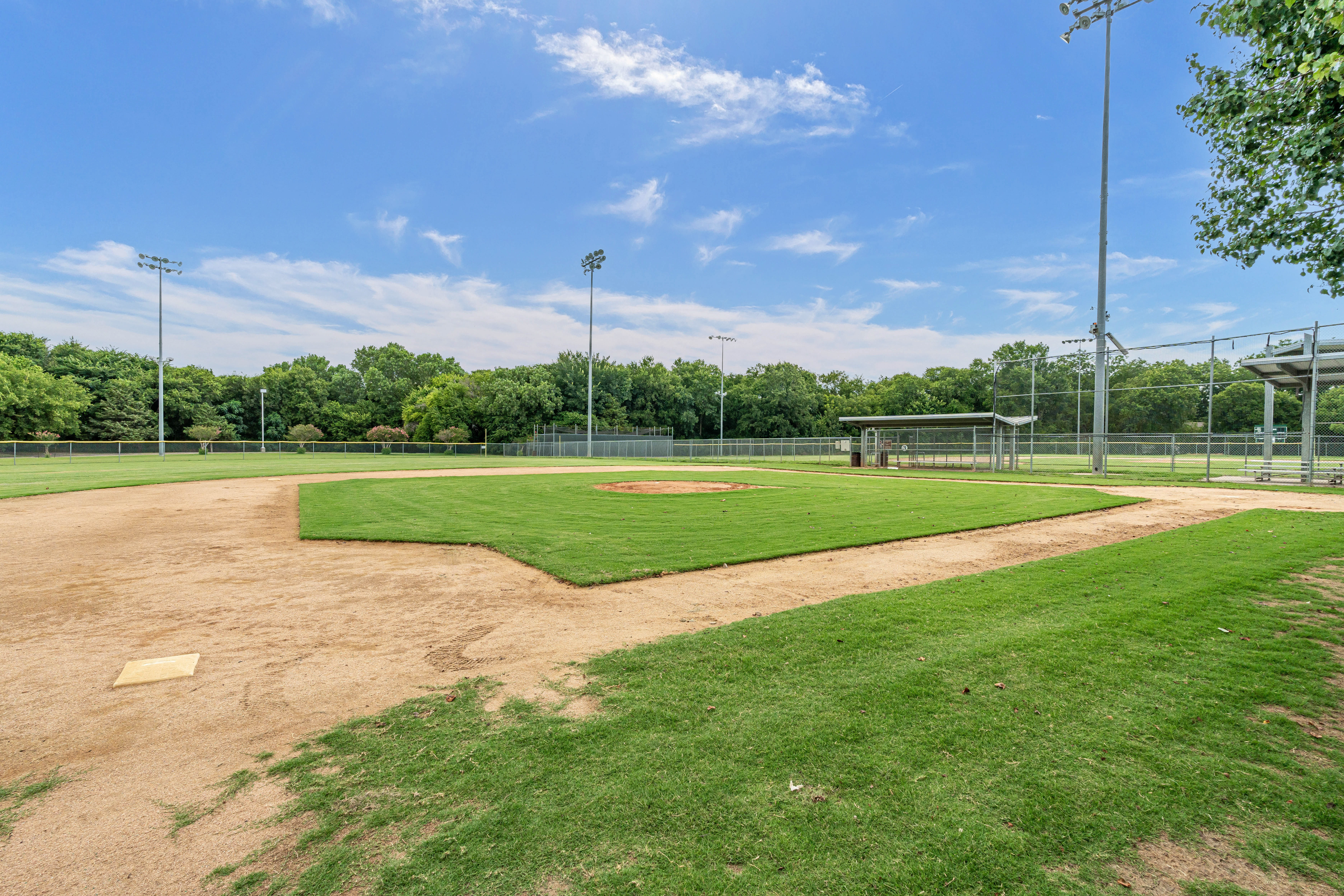 Empty baseball field under a clear blue sky. The diamond's sandy base paths meet lush green grass, flanked by tall lights and a dugout. Peaceful vibe.
