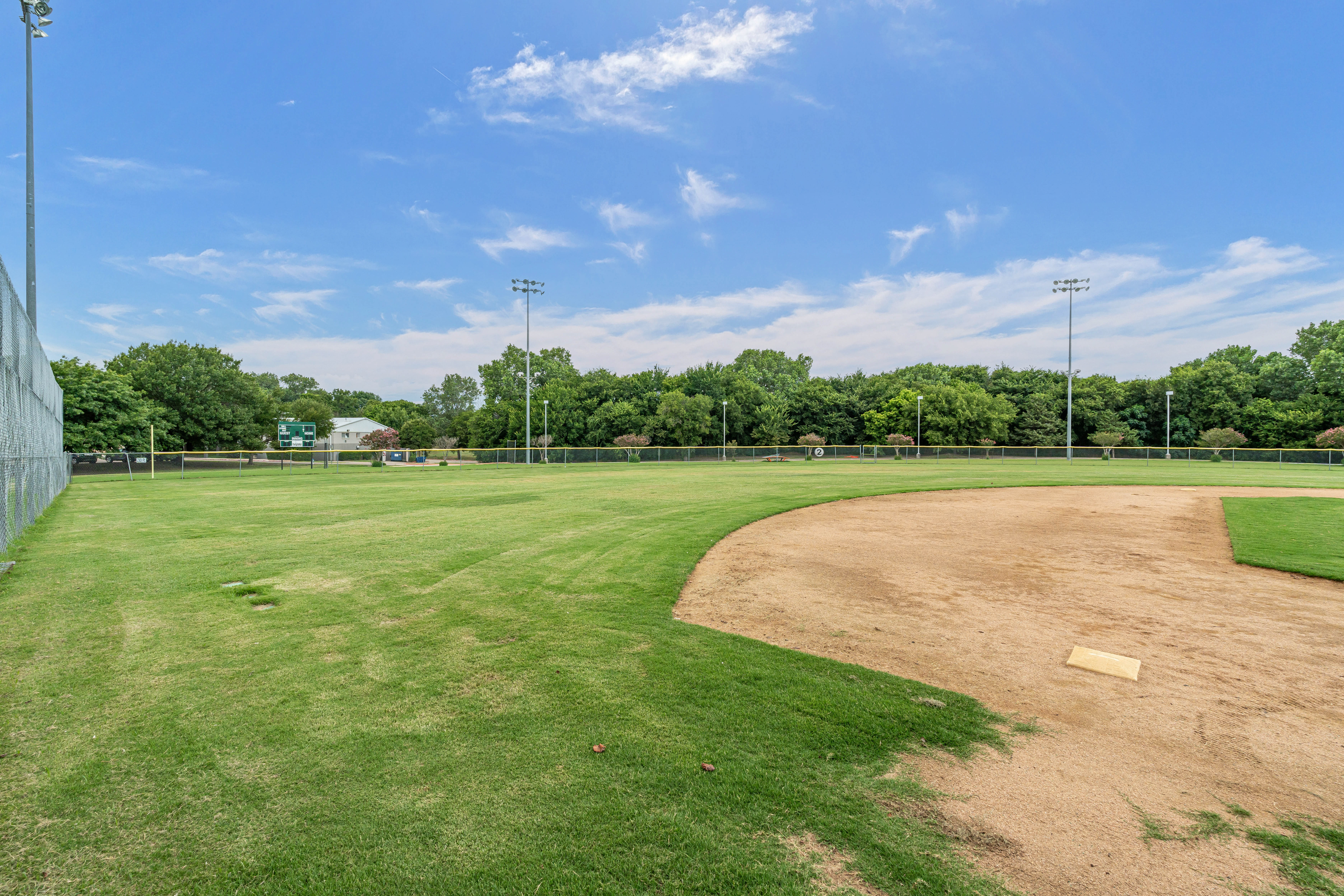 A baseball field with green grass, a dirt infield, a base, and a scoreboard in the distance, surrounded by trees under a blue sky.
