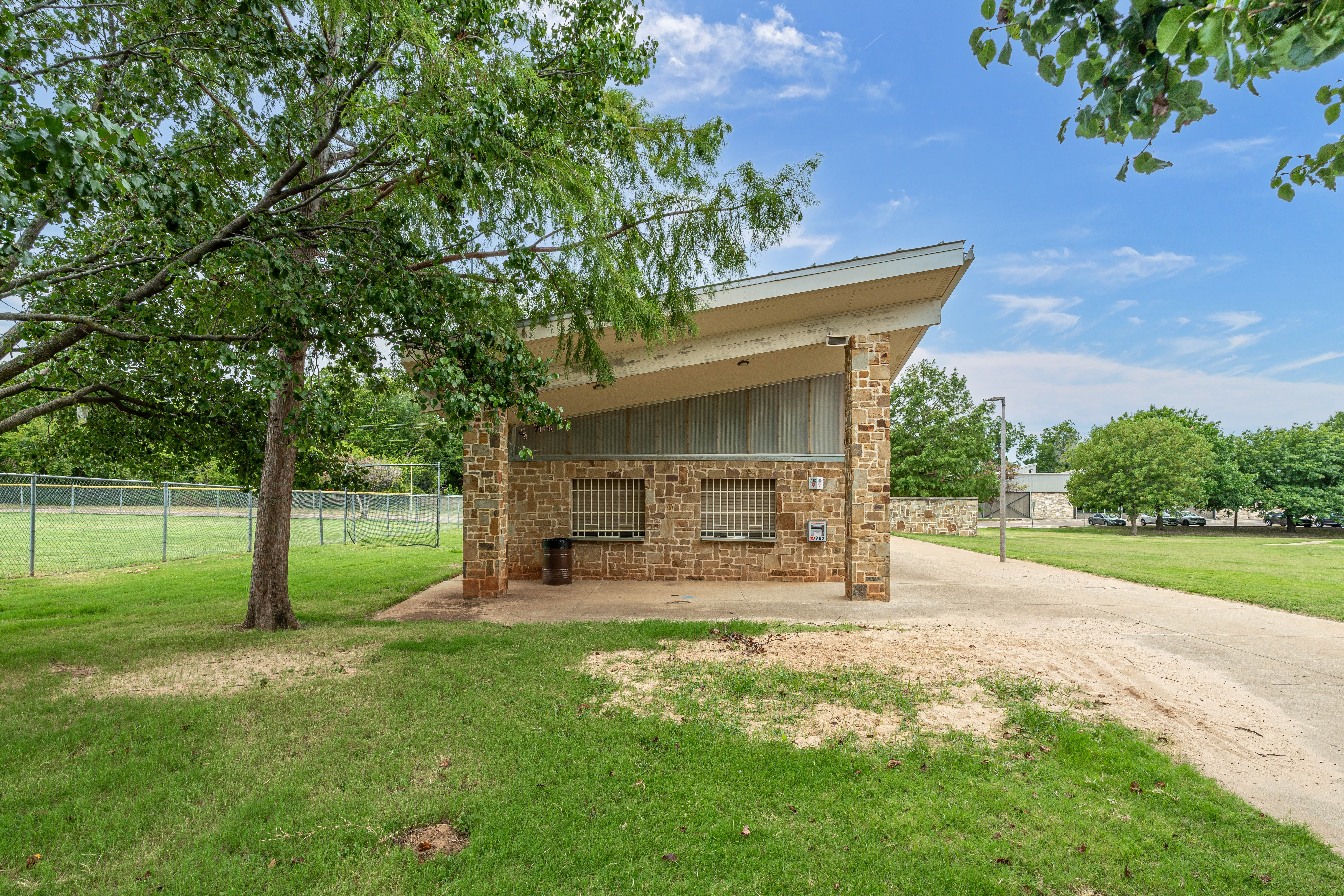A small stone building with barred windows, set in a green park with trees. A dirt pathway leads up to the structure under a clear blue sky.
