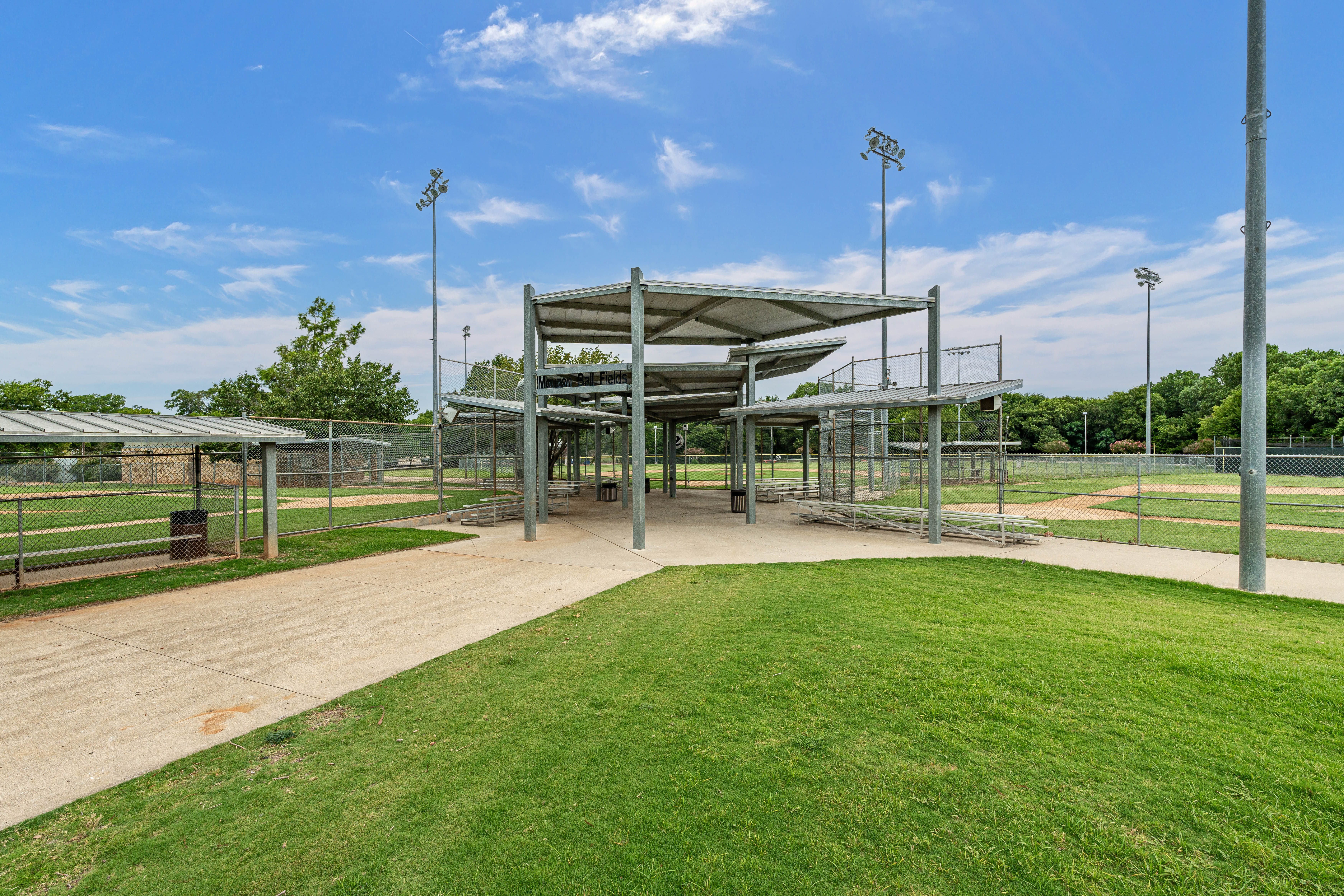 Covered central seating and walkway area between multiple baseball fields, with bleachers and tall light poles under a clear sky.