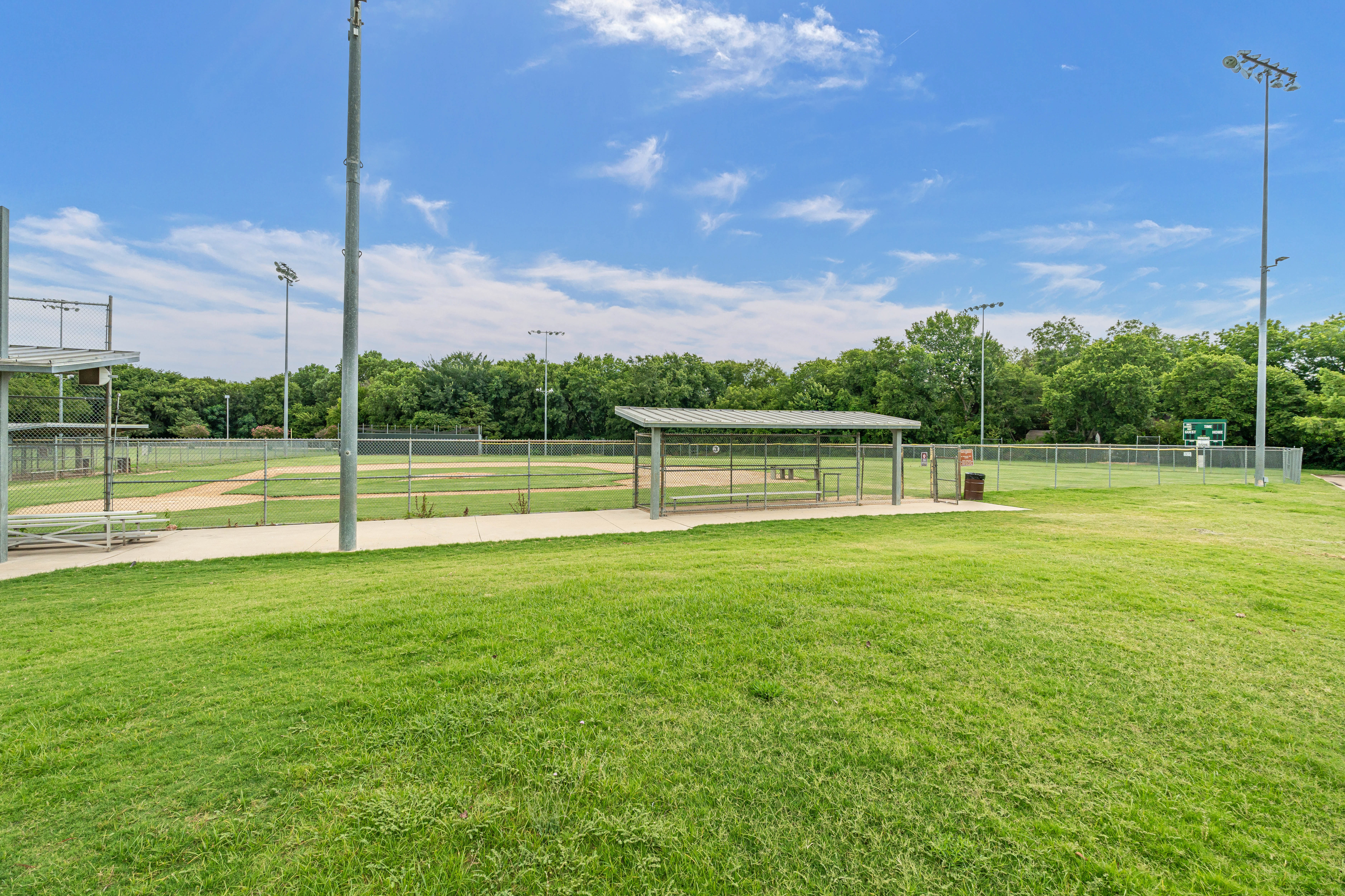 Empty baseball field with a covered dugout, surrounded by green grass and tall light poles under a blue sky.