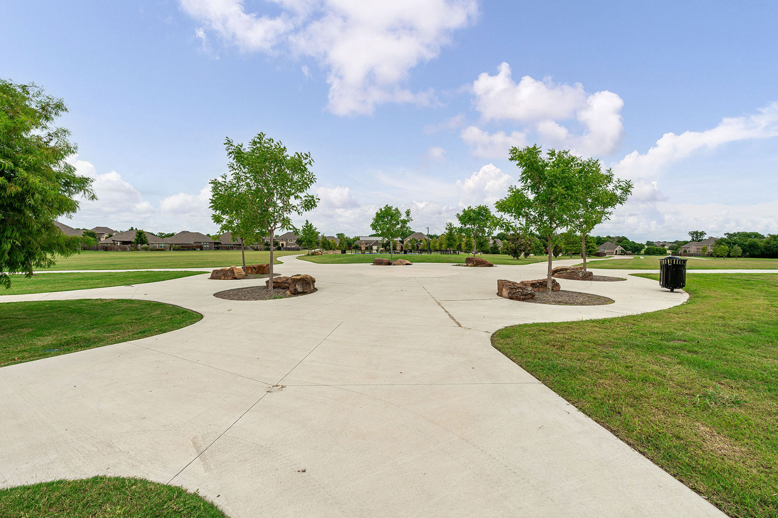 A wide concrete pathway with trees and rocks runs through a green park under a partly cloudy sky, with houses visible in the background.