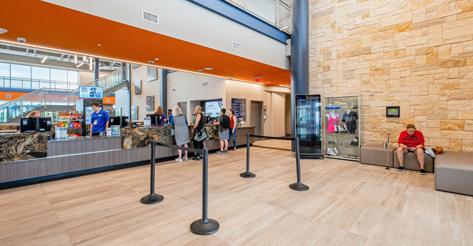 Modern lobby with people standing in line at a counter, and one person sitting on a bench against a stone accent wall.