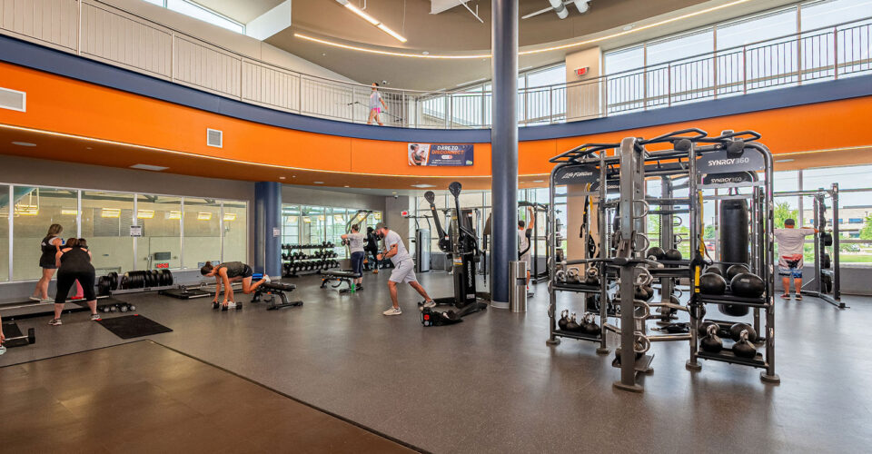 Modern gym with exercise equipment, weights, and people working out in a spacious, well-lit facility with large windows and an upper observation area.