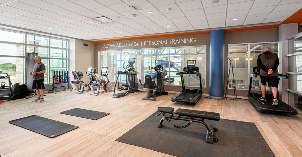 A gym room with treadmills, exercise bikes, mats, a bench, and two people working out; signage reads "Active Adults 60+ | Personal Training.