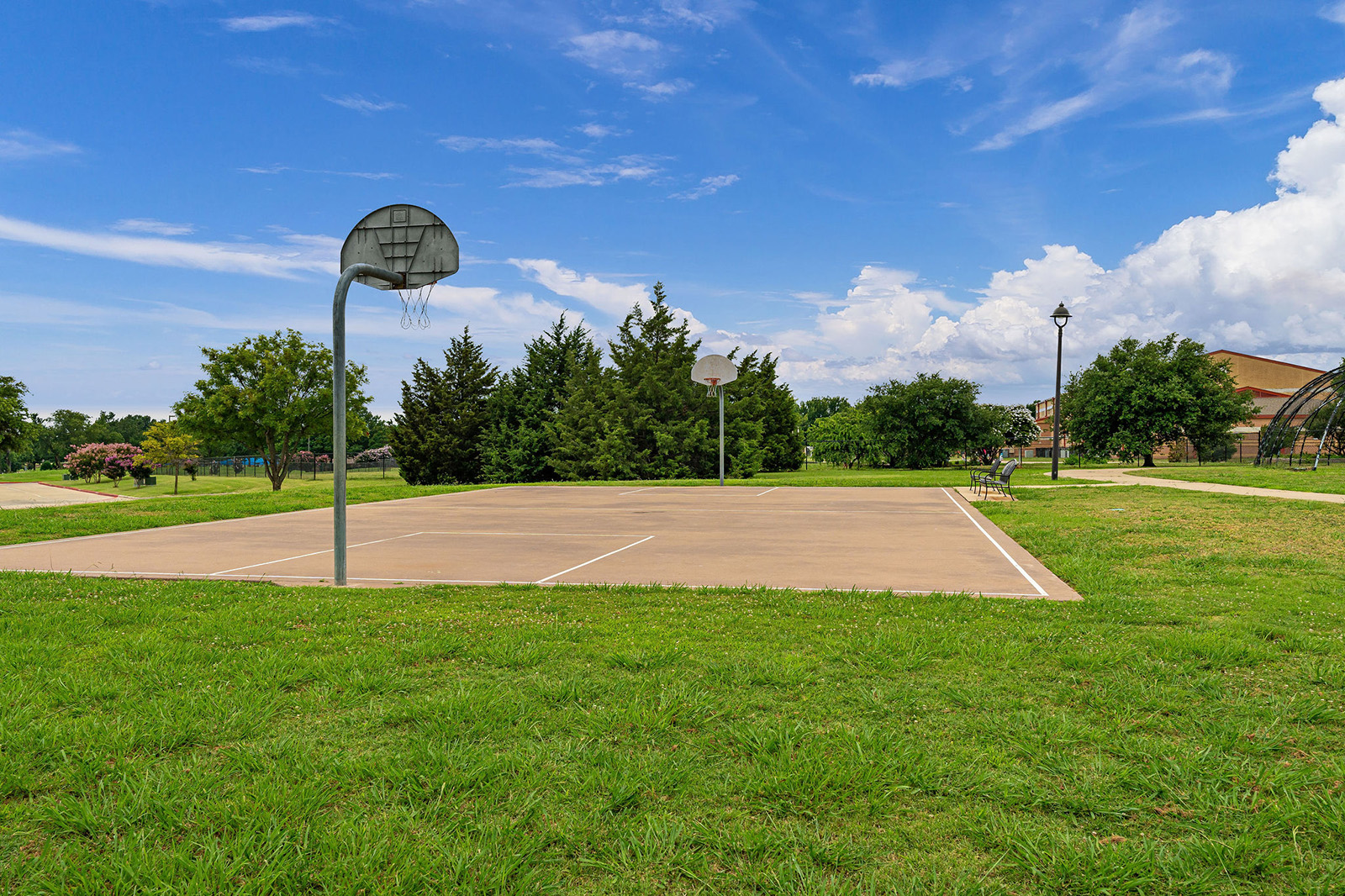 Outdoor basketball court with two hoops, surrounded by grass and trees under a partly cloudy blue sky.