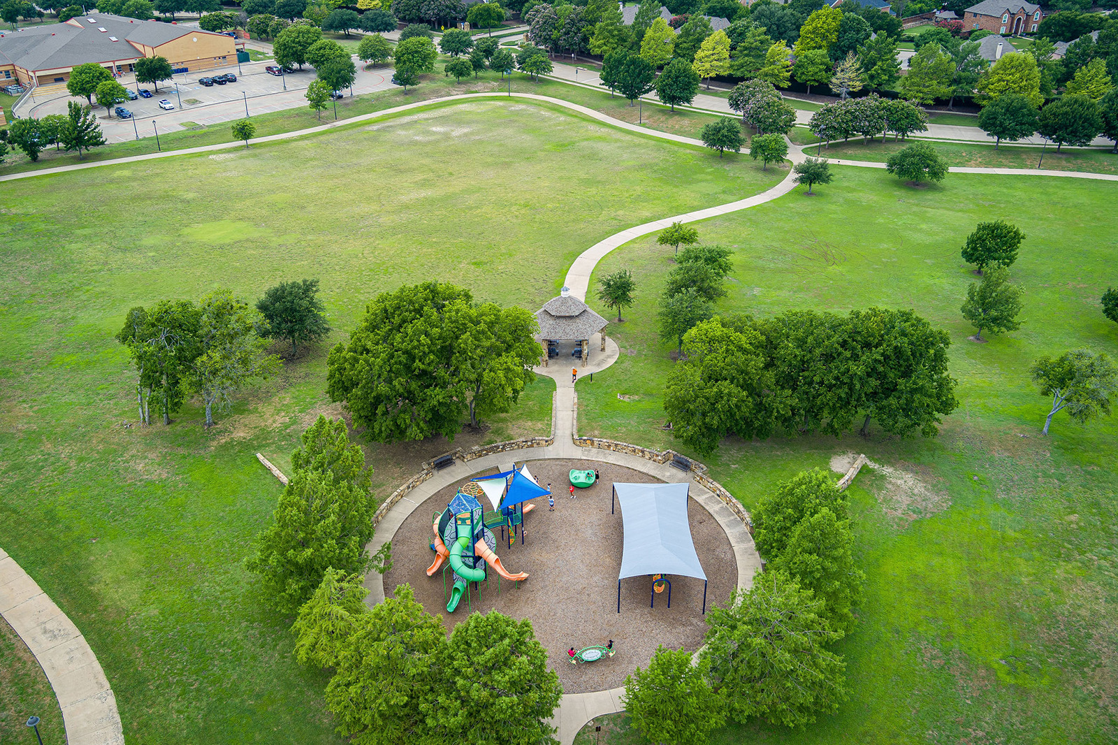 Aerial view of a park with a playground, canopy-covered seating area, walking paths, green lawns, and scattered trees. Nearby buildings and a parking lot are visible in the background.
