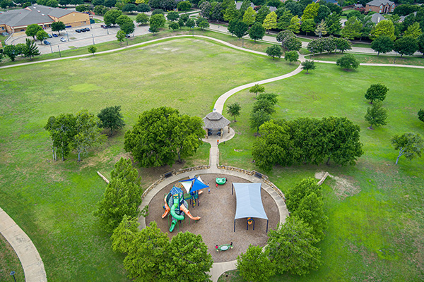 Aerial view of a park with a playground, shaded seating areas, walking paths, trees, and a nearby parking lot.