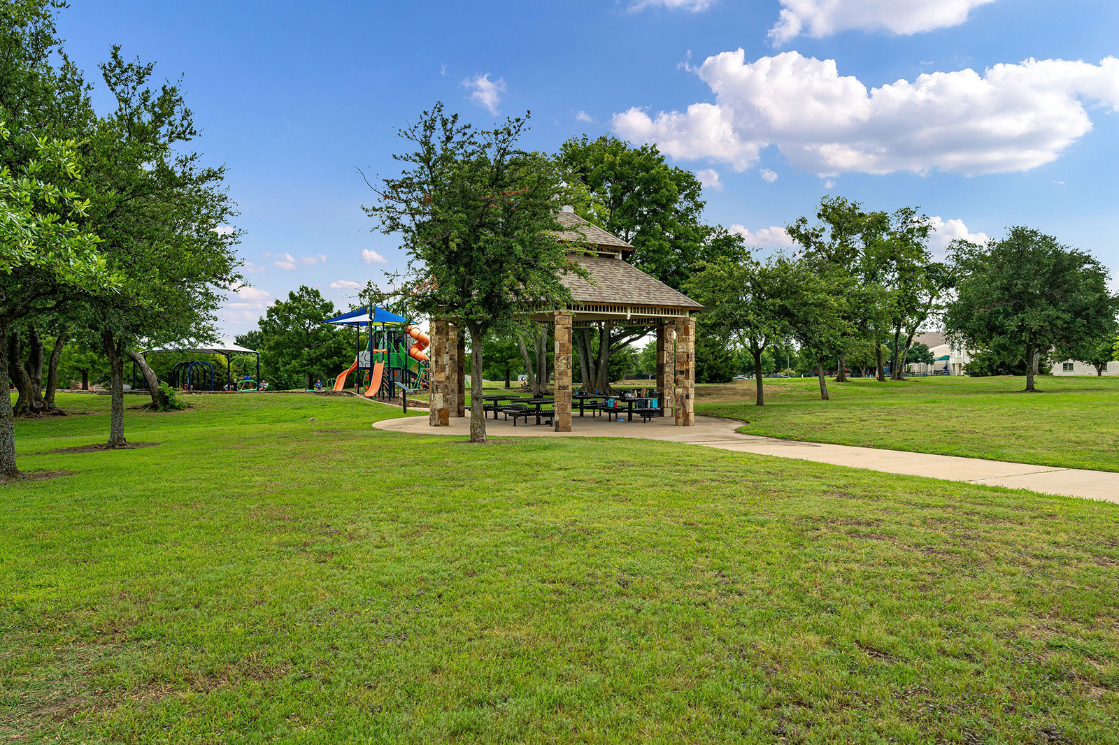 A park with a stone pavilion, picnic tables, trees, a playground with slides, and a paved walkway under a partly cloudy sky.