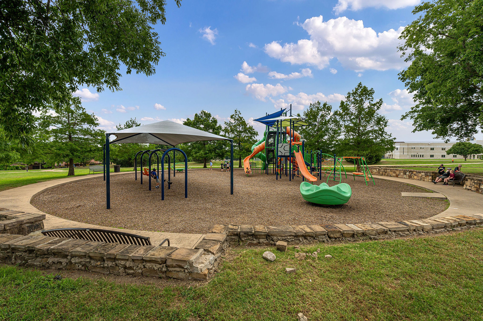 A playground with slides, swings, climbing structures, and a shaded area, surrounded by trees and a stone border on a sunny day.