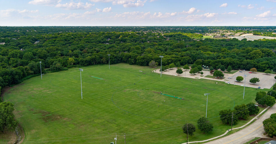 Aerial view of a large, empty grass sports field surrounded by trees, with parking lots and light poles visible, under a blue sky with scattered clouds.
