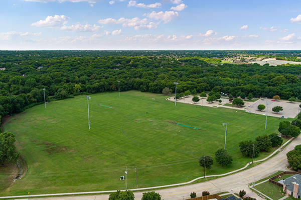 Aerial view of a large, empty grassy sports field surrounded by trees, parking lots, and a few light poles under a partly cloudy sky.