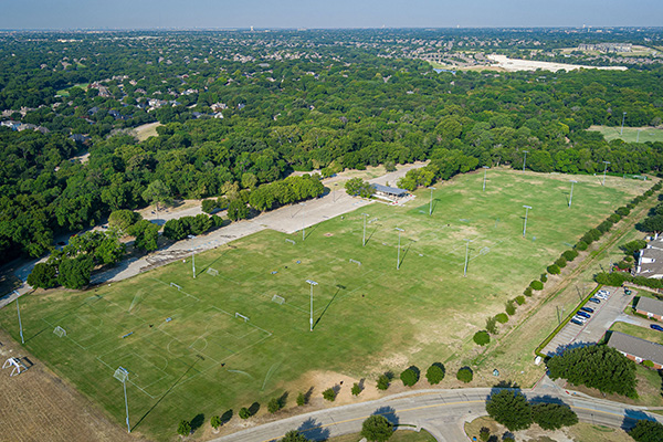 Aerial view of large outdoor sports fields with multiple soccer goals, surrounded by trees and nearby buildings under a clear sky.