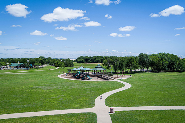 A large green park with a playground under shaded structures, surrounded by paths and trees, beneath a blue sky with scattered clouds.