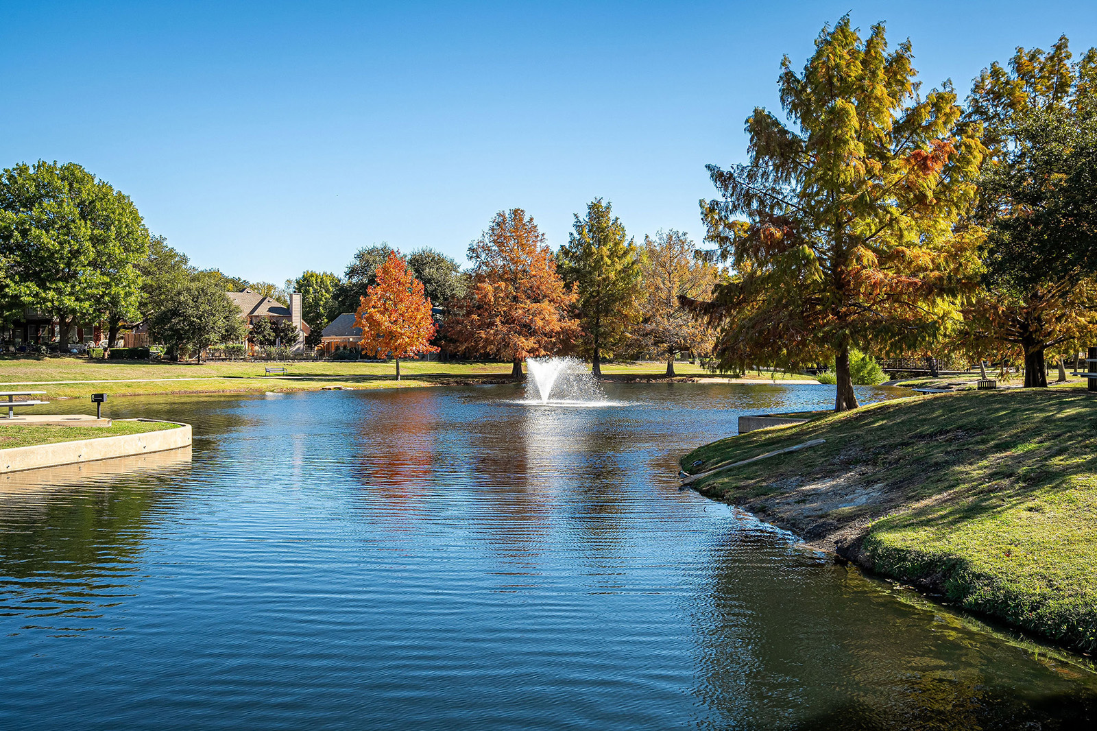 A small pond with a fountain in the center, surrounded by trees with autumn foliage and grassy areas under a clear blue sky.