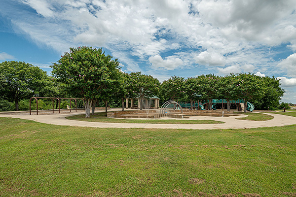 A circular splash pad with water features is surrounded by green grass, trees, and a partly cloudy sky in a park.