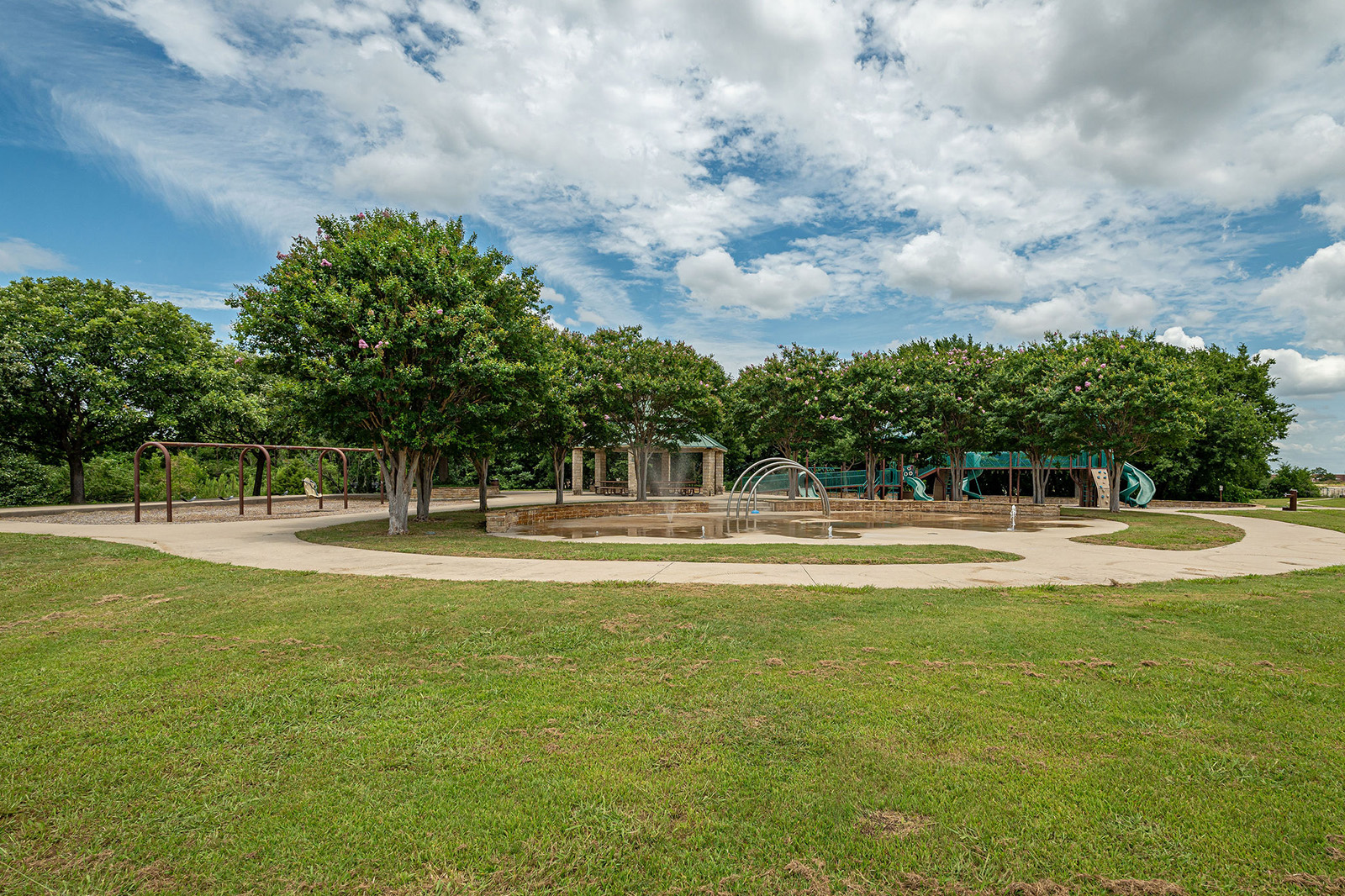 A circular splash pad with fountains is surrounded by trees, a playground, and swings under a partly cloudy sky in a grassy park.