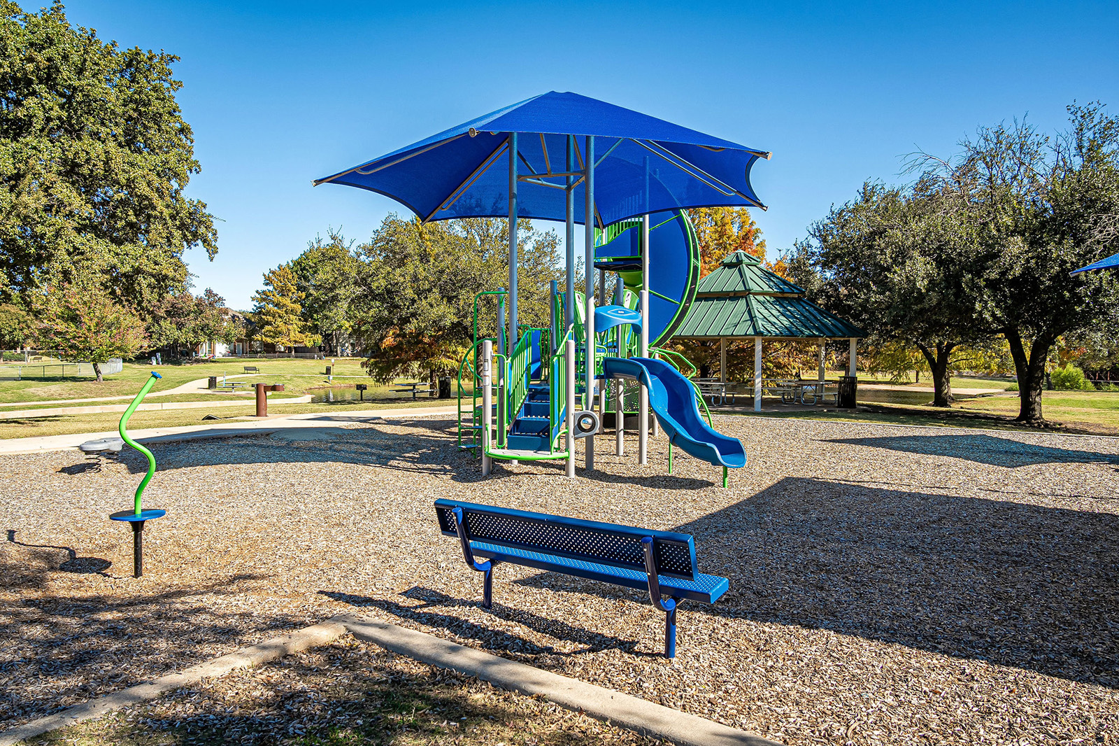 A playground with a blue play structure, slides, benches, and a large shade canopy, surrounded by trees and a grassy park area on a sunny day.