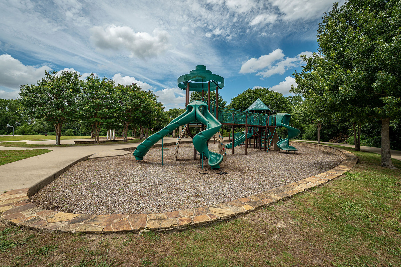 A playground with multiple green slides, climbing structures, and a gravel surface, surrounded by trees and a stone border under a partly cloudy sky.