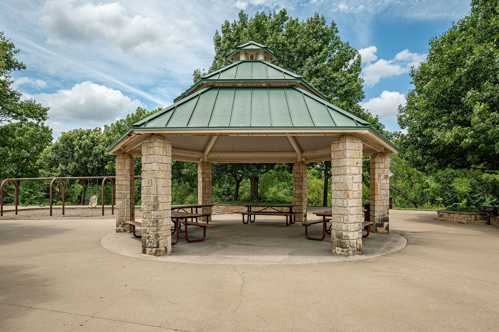 Hexagonal stone pavilion with a green metal roof, picnic tables underneath, and a playground structure in the background, surrounded by trees.
