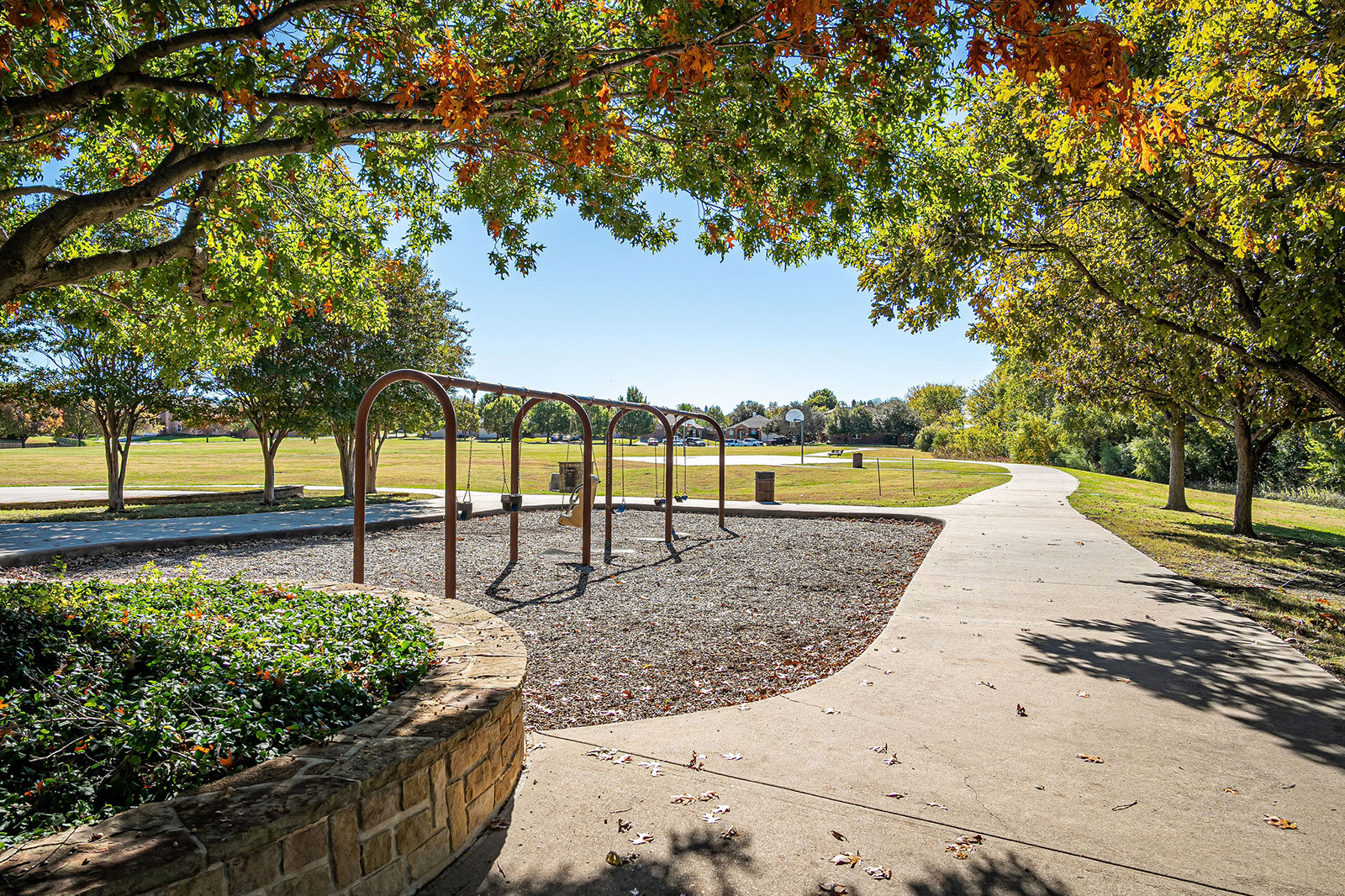 A paved walking path curves past empty swing sets and trees in a sunny park with green grass and scattered autumn leaves.