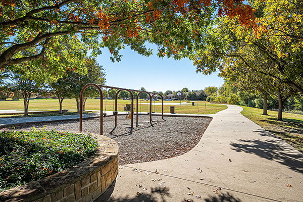 A playground with empty swings is next to a curved pathway in a park with trees and open grassy areas on a sunny day.