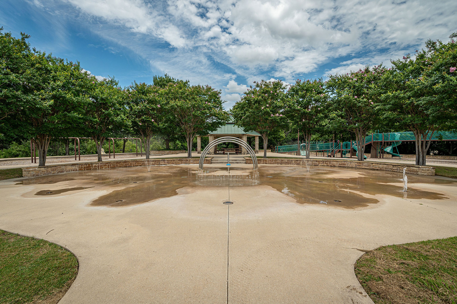 Concrete splash pad with metal arches and small fountains, surrounded by trees and a playground structure in the background under a partly cloudy sky.