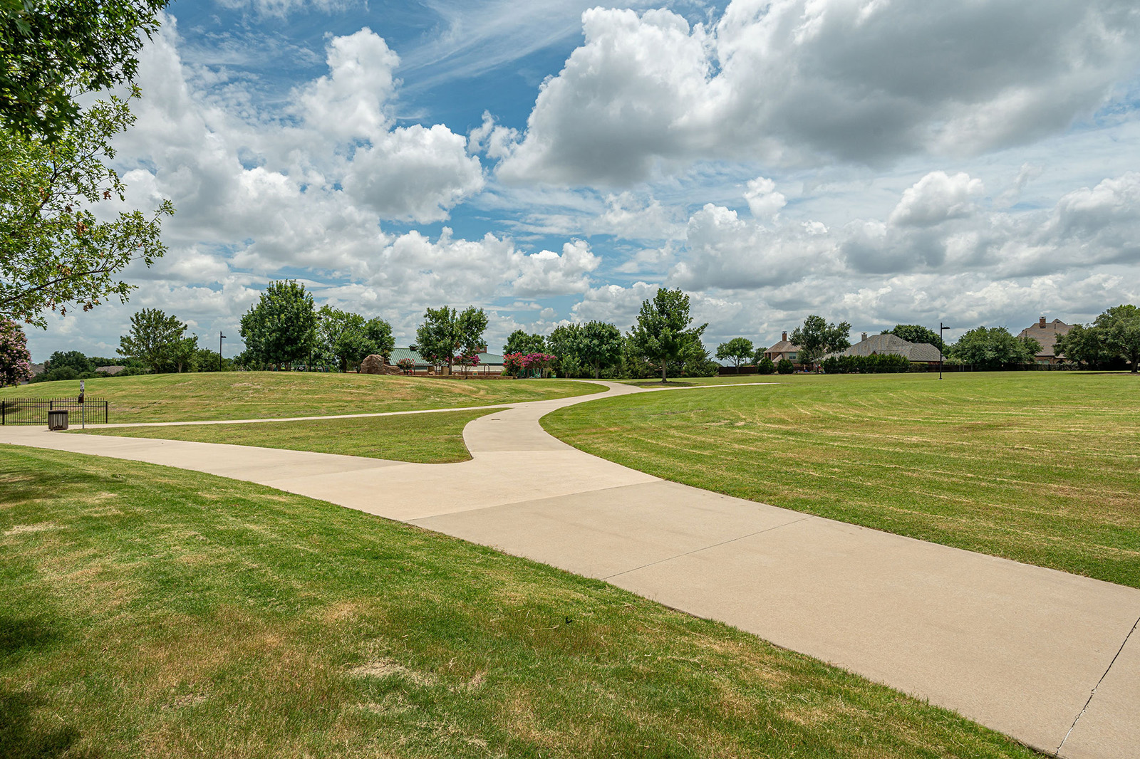 A concrete walking path curves through a grassy park under a partly cloudy sky, with trees and houses in the background.
