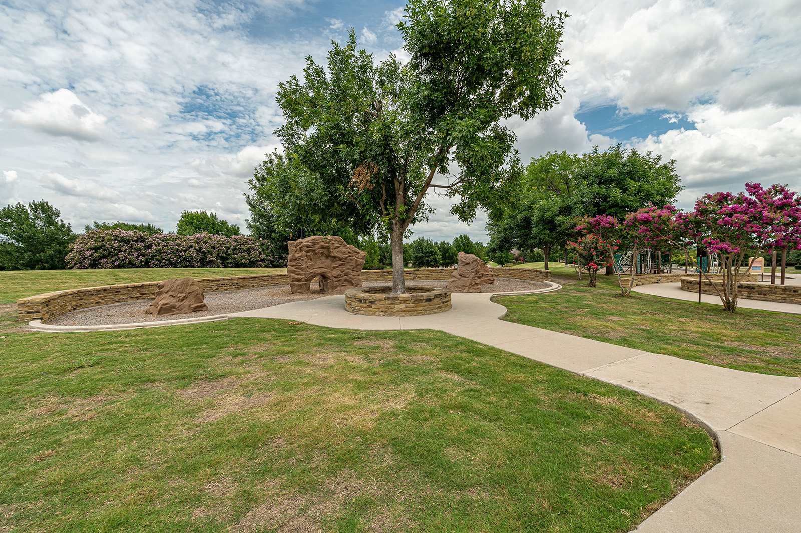 A park scene with a concrete walkway, grassy areas, a central tree, rock formations, and blooming shrubs under a partly cloudy sky.