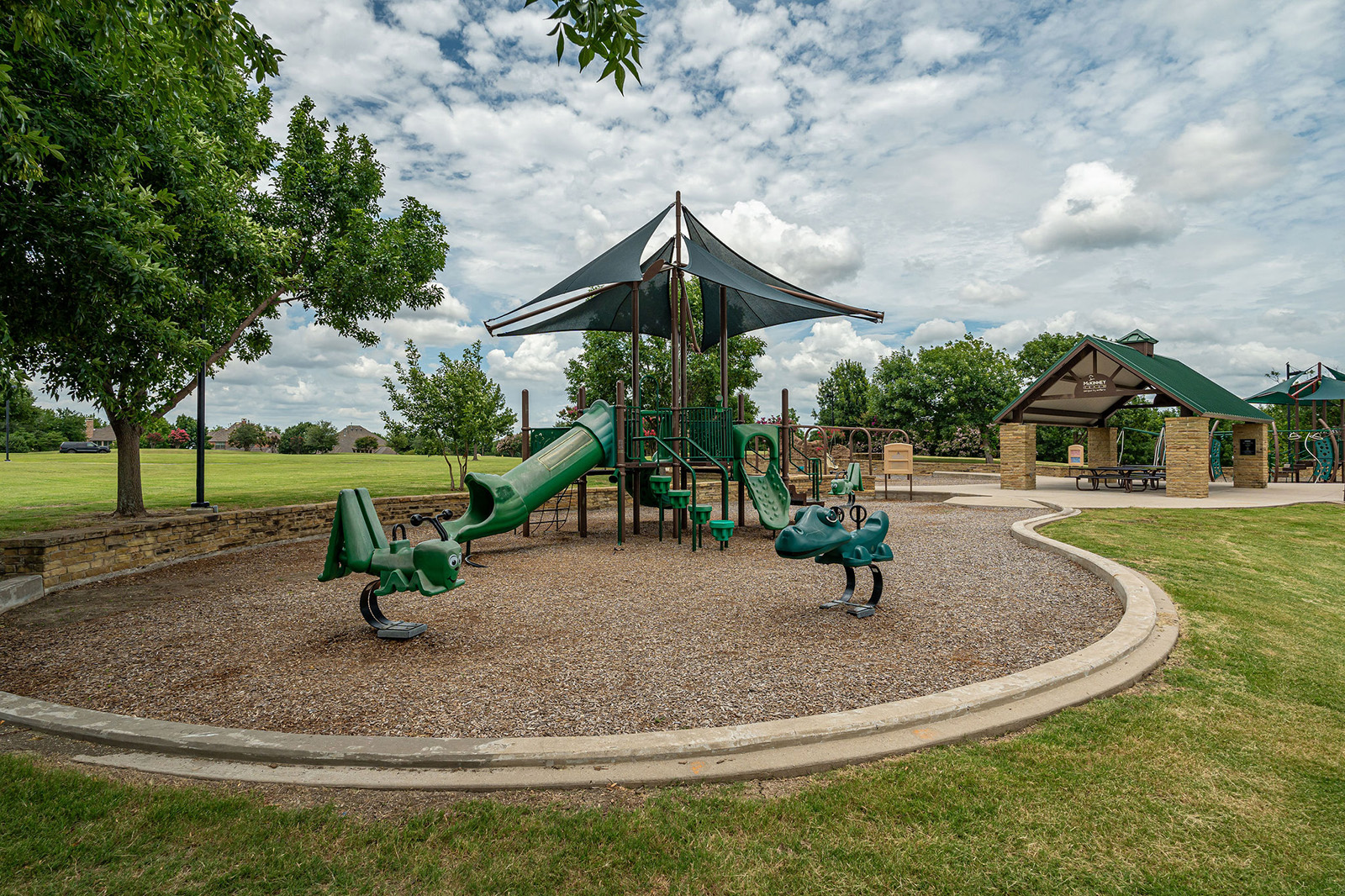 Playground with green equipment and shade structures, surrounded by grass and trees, under a partly cloudy sky. Picnic pavilion visible in the background.